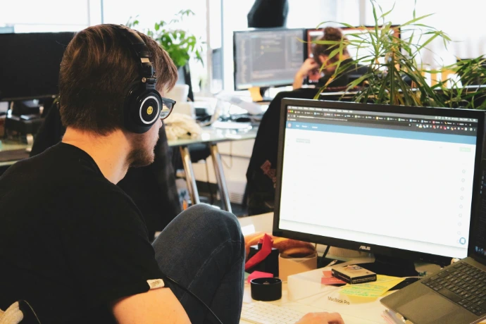 Un programador joven con auriculares grandes negros trabajando concentrado frente al monitor de su ordenador en una oficina compartida con otros compañeros al fondo.
