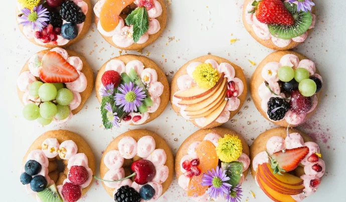 Vista cenital de variedad de doce dónuts organizados en cuadrícula sobre una superficie blanca. Están elaboradamente decorados con glaseado rosa, frutas frescas (fresas, uvas, moras, kiwi, melocotón) y flores comestibles.